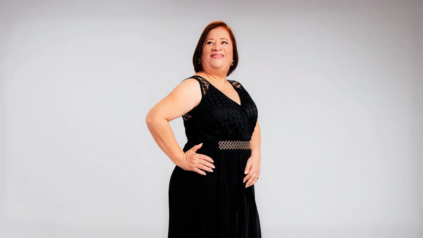 Thyna Ramos sorri olhando para o alto, de vestido preto, em um estúdio de fundo claro