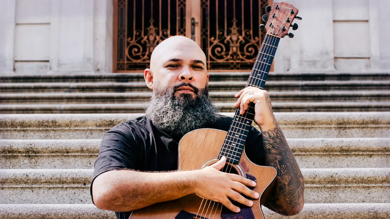 Vitor Teixeira abraça o violão diante da escadaria e das portas de ferro de uma igreja